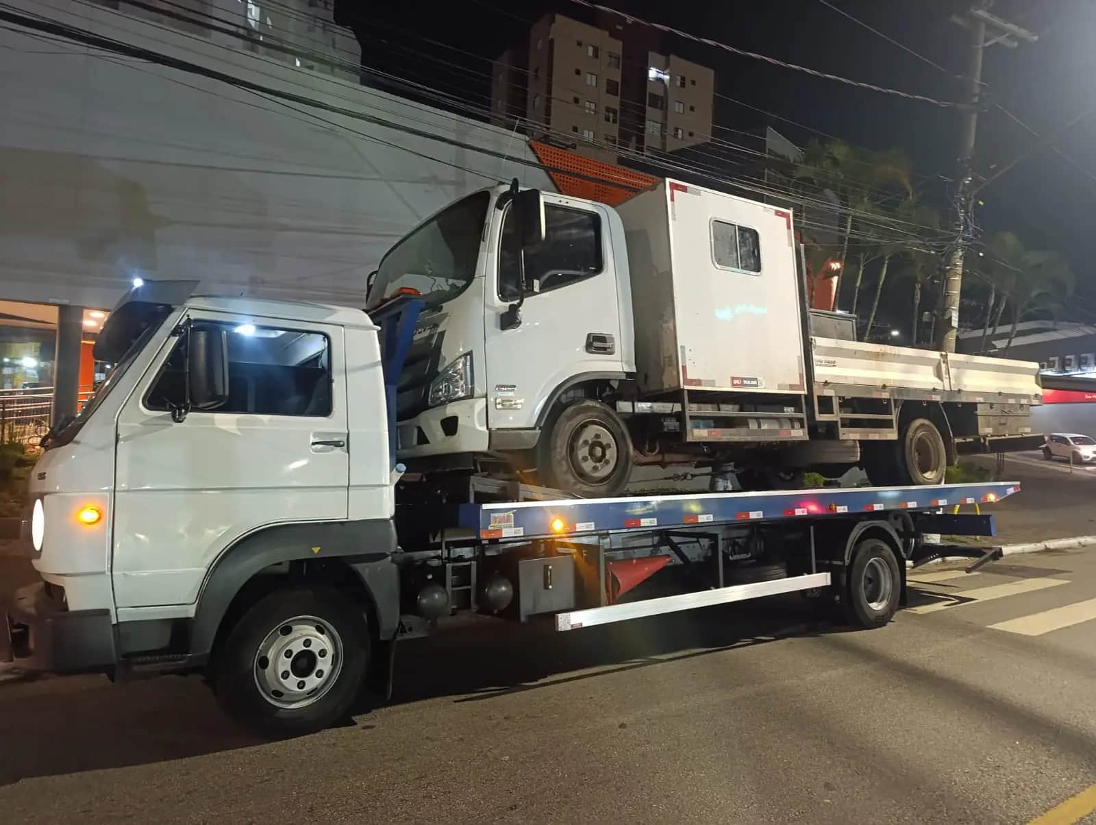 Caminhão utilitário carregado na plataforma do guincho da JHC em avenida iluminada, durante a noite.