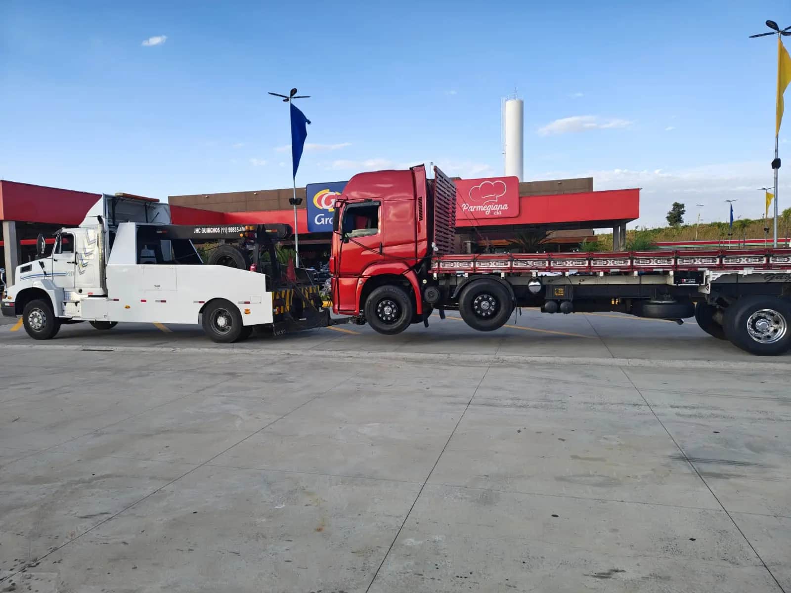 Guincho pesado da JHC acoplado a um cavalo mecânico vermelho com carreta carregada, visto de lado no pátio de um posto.