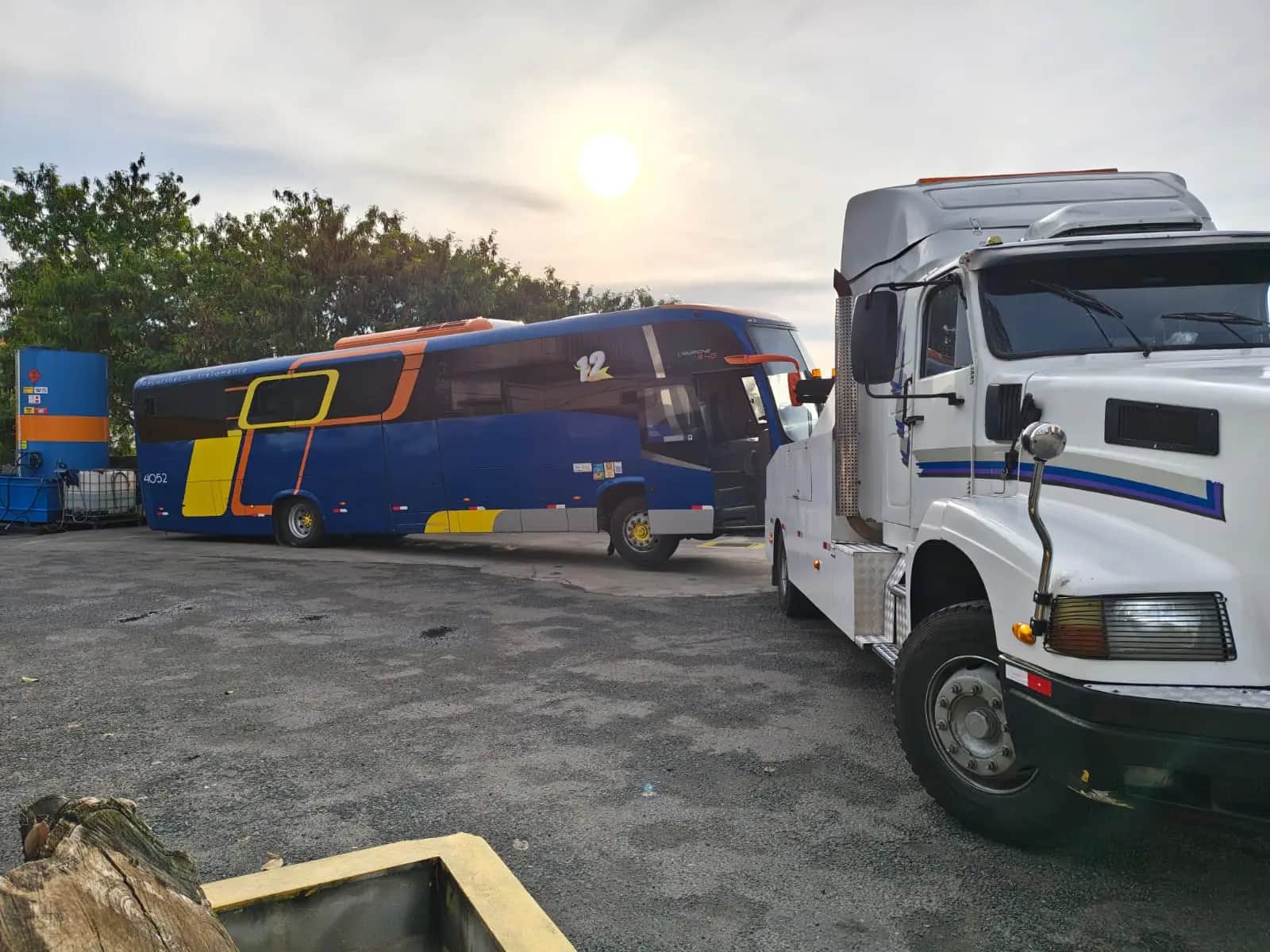 Ônibus rodoviário azul e amarelo ao lado do guincho pesado da JHC em um pátio, no fim da tarde.
